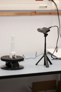 Przyrząd przypominający lampkę biurową skierowany na przedmiot przypominający zabawkową wieżyczkę stojący na małym okrągłym podeście.