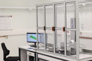 W przezroczystej gablocie widoczny przyrząd o nazwie profilometr stykowy. Na dalszym planie monitor na blacie stołu.