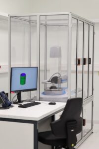 Na stole z białym blatem stoi monitor. Obok fotel biurowy. Na dalszym planie przezroczysta komora z przyrządem do pomiaru.