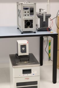 Na niedużym stole dwa przyrządy pomiarowe, pod nim biała skrzynka.