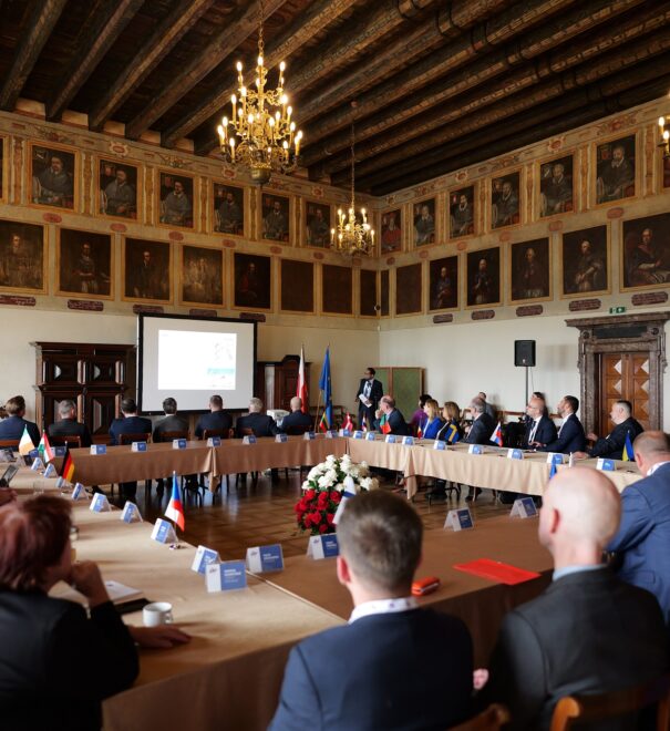 W pięknej, starej sali, z drewnianą podłogą i sufitem, przy stołach ustawionych w kształcie kwadratu siedzą elegancko ubrani uczestnicy spotkania. W głębi sali widać monitor wyświetlającym prezentację.