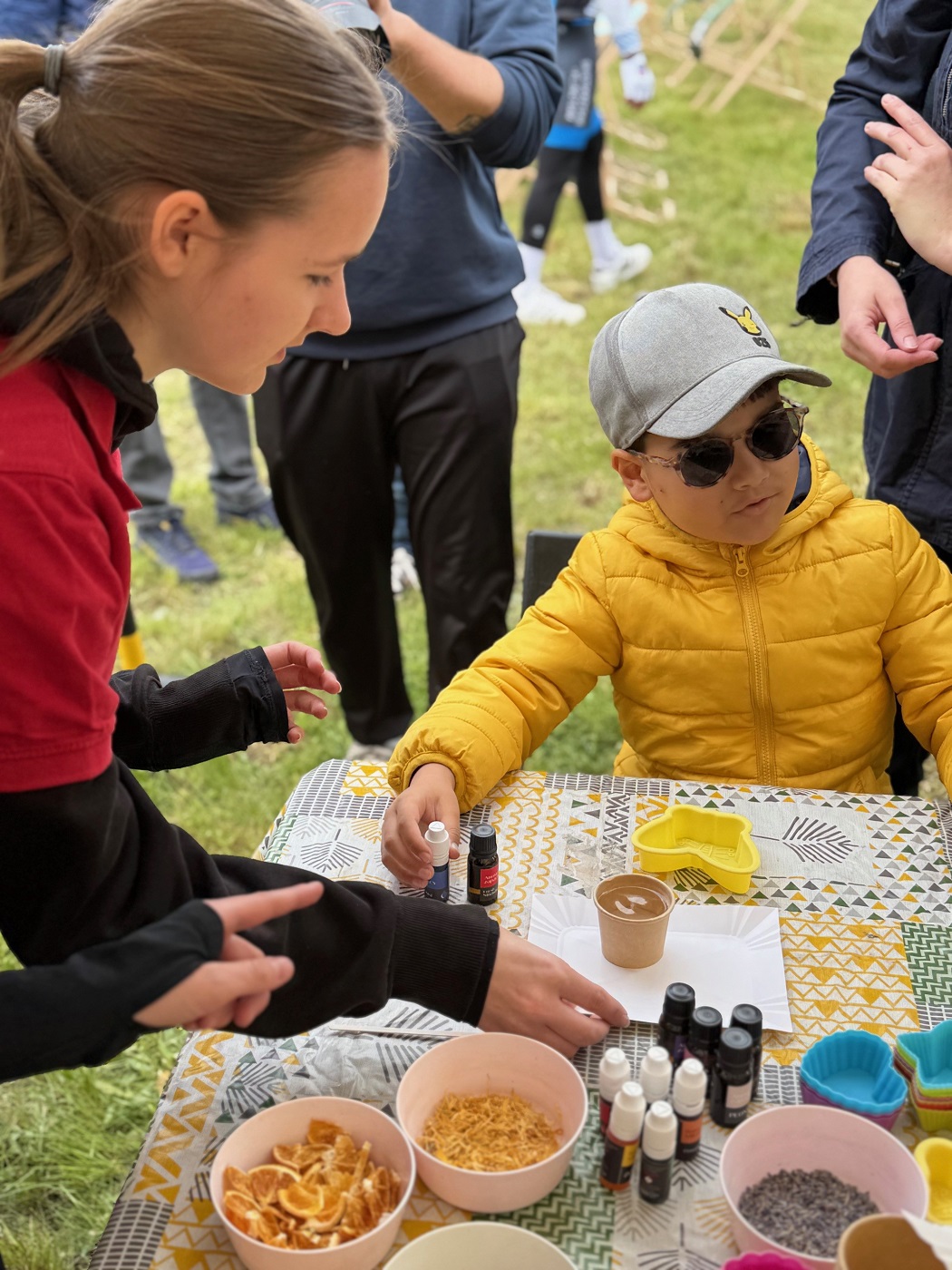 Chłopak w żółtej kurtce, przyciemnianych okularach i czapce z daszkiem stoi przy stoliku, na którym leżą różne przedmioty przydatne do zabawy i metrologicznych doświadczeń.