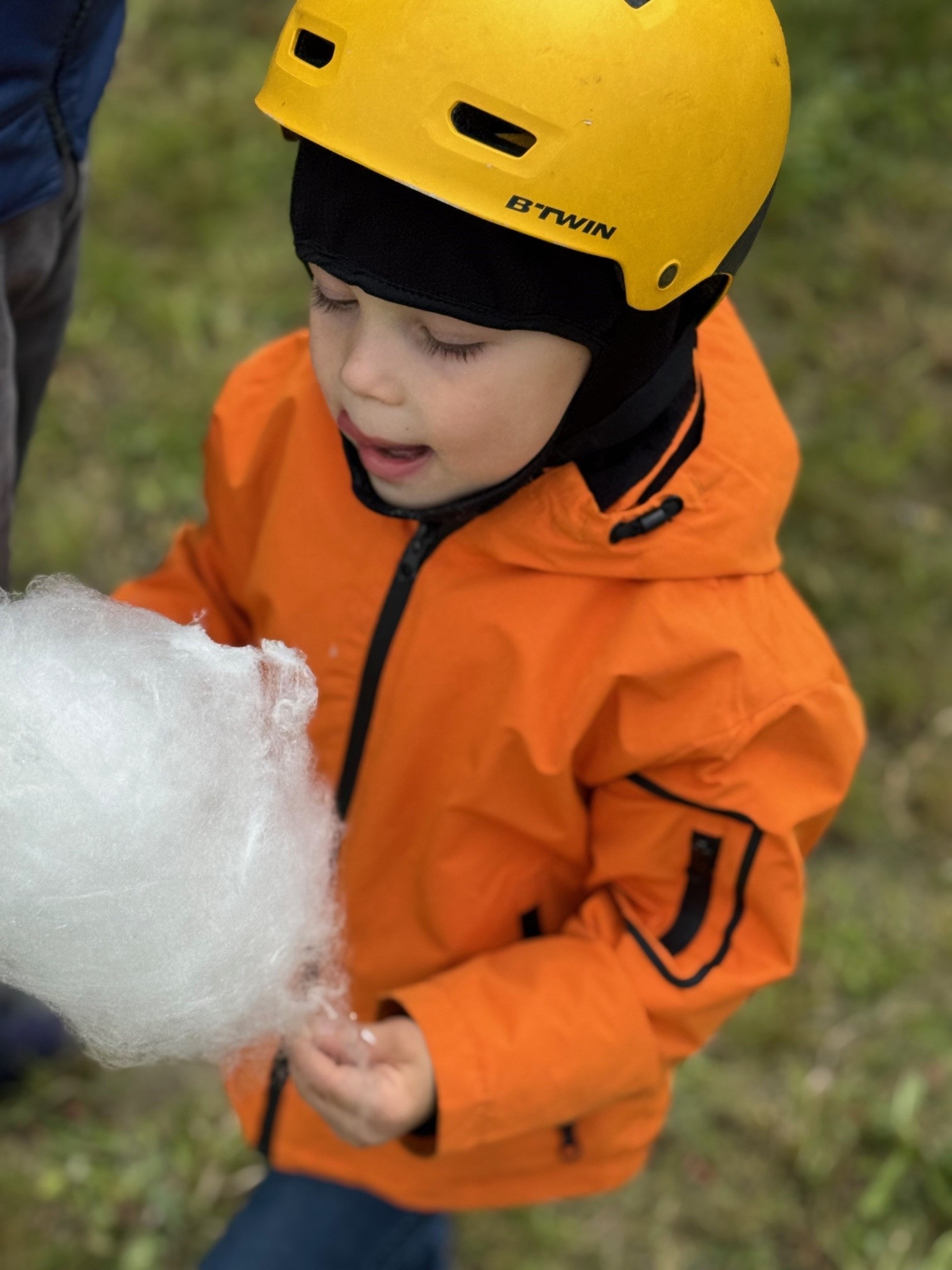 Dziecko w pomarańczowym kombinezonie i żółtym kasku trzyma w ręku watę cukrową na patyku.