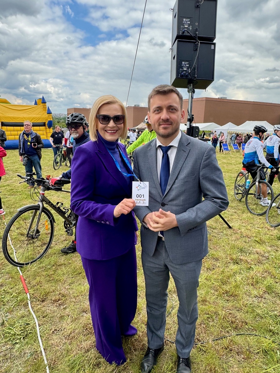 Mężczyzna w garniturze i kobieta w niebieskim kombinezonie pozują do zdjęcia. W tle widać dzieci i młodzież na rowerach, na trawie.