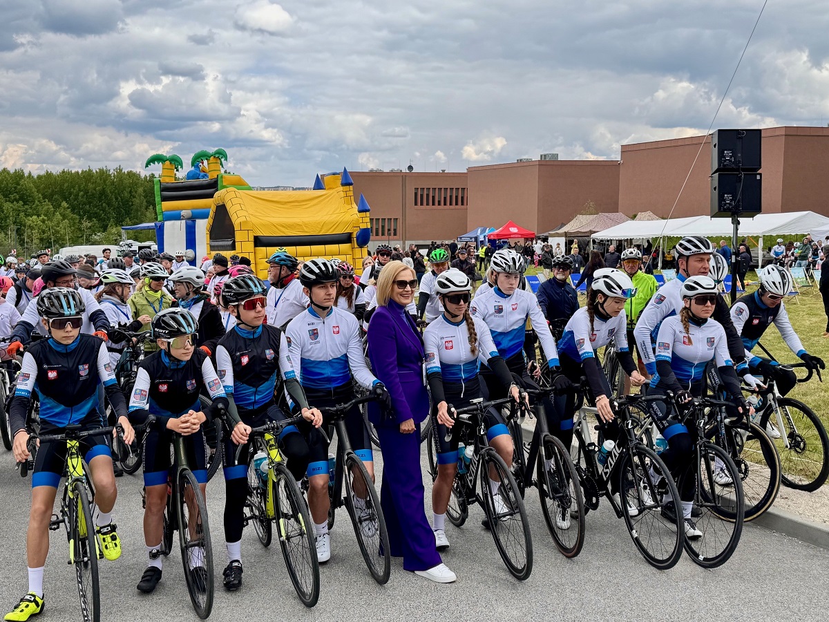 Młodzi kolarze w strojach kolarskich szykują się do startu. Wśród nich stoi kobieta w niebieskim, eleganckim kombinezonie.