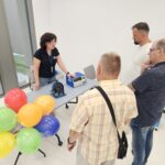 Trzech mężczyzn stoi przy stoliku, na którym leżą dwa przyrządy pomiarowe. Stanowisko obsługuje kobieta w ciemnej bluzce.