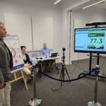 Mężczyzna w szarej marynarce stoi przed monitorem, na którym wyświetlony został pomiar ćwiczenia, w którym wziął udział. Na dalszym planie, Przy ścianie siedzi dwóch mężczyzn, obserwujących pomiar.