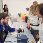 Dwie uśmiechnięte kobiety stoją przy stoisku, na którym przyciskając mały przycisk, można zbadać swój refleks. Za stołem siedzi obsługujący stanowisko mężczyzna.