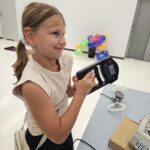 Dziewczynka w jasnej koszulce trzyma w rękach podłużny przedmiot - przyrząd metrologiczny.