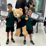 Dwóch jednakowo ubranych, w ciemnozielone koszulki, uśmiechniętych chłopaków w okularach trzyma w rękach dyplomy. Obaj trzymają też dużą maskotkę misia.