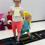 Chłopak i dziewczynka stoją na niedużej czarnej macie z podziałką pokazującą odległość. Dziewczynka trzyma w rękach dyplom.