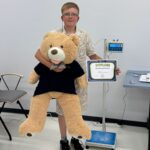 Chłopak w okularach trzyma w jednej ręce dużego misia - maskotkę, w drugiej ręce dyplom.