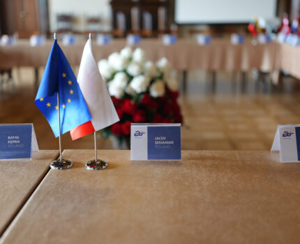 Na pierwszym planie flagi Polski i Unii Europejskiej. na drugim bukiet z białych i czerwonych kwiatów.