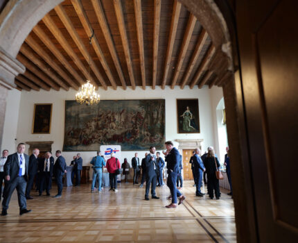 Duża, zabytkowa sala ze starymi obrazami na ścianach, brązową posadzką, po której spacerują uczestnicy spotkania.