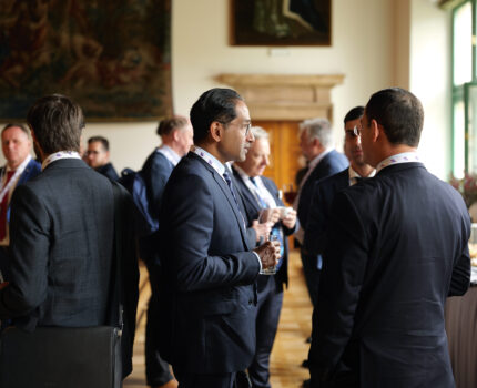 Kilku mężczyzn w ciemnych garniturach rozmawia w zabytkowej sali.