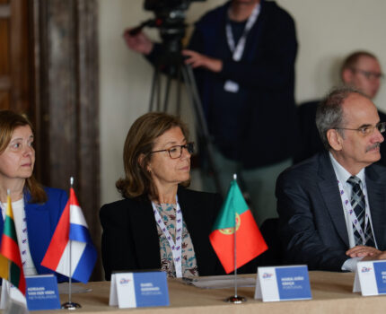 Kobieta w okularach siedzi przy stole, przed nią ustawiona flaga Portugalii i wizytówka.