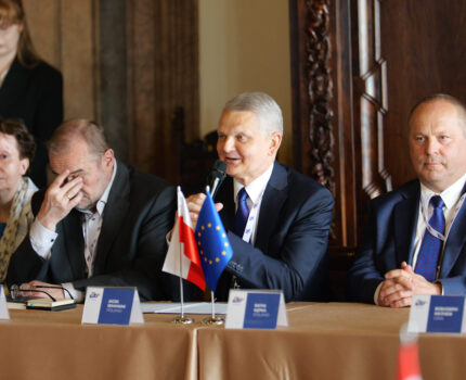 Trzech mężczyzn w garniturach oraz jedna kobieta siedzą przy stole, na którym stoją flagi polska i Unii Europejskiej. Mężczyzna w środku mówi do mikrofonu, który trzyma w ręku.