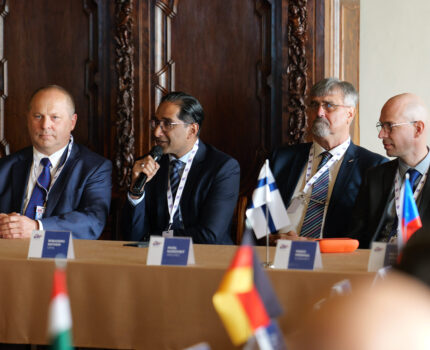 Czterech mężczyzn w garniturach siedzi przy stole, na którym stoi flaga Finlandii oraz wizytowniki. Mężczyzna w środku mówi do mikrofonu, który trzyma w ręku.