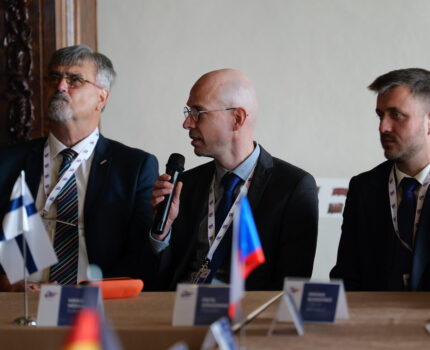 Trzech mężczyzn w garniturach siedzi przy stole. Łysawy Mężczyzna w okularach w środku mówi do mikrofonu, który trzyma w ręku.