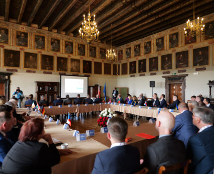 W pięknej, starej sali, z drewnianą podłogą i sufitem, przy stołach ustawionych w kształcie kwadratu siedzą elegancko ubrani uczestnicy spotkania. W głębi sali widać monitor wyświetlającym prezentację.