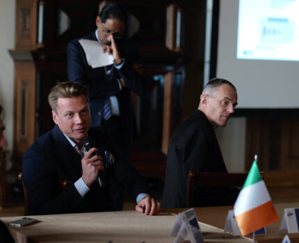 Kilku uczestników spotkania siedzi przy stole. Jeden z nich ma w ręku mikrofon.