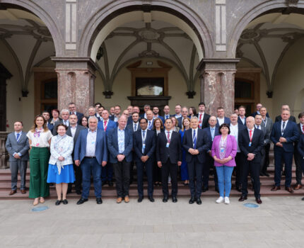 Grupa kilkudziesięciu osób, uczestników spotkania pozuje do zdjęcia przed budynkiem z arkadami.