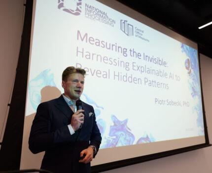 Mężczyzna w ciemnym garniturze podczas prezentacji. Za nim wyświetlona prezentacja.