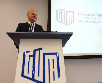 Mężczyzna w ciemnym garniturze stoi przy mównicy z logotypem Głównego Urzędu Miar. Przed nim mikrofon.