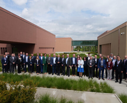 Grupa ponad dwudziestu osób pozuje do zdjęcia na terenie Kampusu GUM. Stoją w alejce pomiędzy trawnikami. Obok budynki Kampusu.