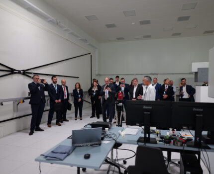 Kilkanaście osób stoi w pomieszczeniu laboratoryjnym, w którym znajdują się przyrządy pomiarowe.