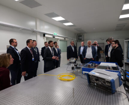 Grupa osób stoi w pomieszczeniu laboratoryjnym, w którym znajdują się przyrządy pomiarowe.