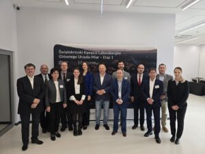 Grupa trzynastu osób pozuje do zdjęcia na tle fototapety z makietą Kampusu GUM.