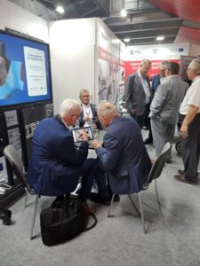 W hali targowej, na jednym ze stoisk kilku mężczyzn siedzi przy niedużym stoliku, a kilku innych stoi i rozmawia. W tle Widoczny ekran wyświetlający materiał filmowy.