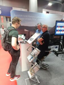 W hali targowej gość podchodzi do stoiska, przy którym siedzi dwóch mężczyzn. Widać rozłożone materiały reklamowe.