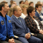 Na pierwszym planie dwóch mężczyzn i kobieta siedzą w jednym rzędzie na sali wypełnionej ludźmi.