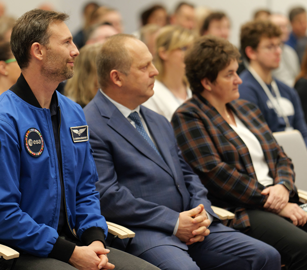 Na pierwszym planie dwóch mężczyzn i kobieta siedzą w jednym rzędzie na sali wypełnionej ludźmi.