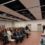 Na dużej sali, w kilku rzędach siedzi grupa kilkudziesięciu osób, które spoglądają na stojących przed nimi kilkoro ludzi.