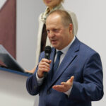 Mężczyzna w średnim wieku, w ciemnoniebieskim garniturze mówi do mikrofonu, trzymanego w ręce. Za nim widoczna uśmiechnięta kobieta.