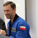 Uśmiechnięty mężczyzna w niebieskiej kurtce z naszytą na ramieniu polską flagą, mówi do mikrofonu.