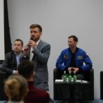 Mężczyzna w szarej marynarce, trzymający mikrofon, zwraca się do kilku osób, siedzących tyłem do zdjęcia. Za mężczyzną z mikrofonem, na fotelach siedzi dwóch mężczyzn.