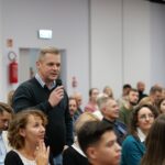 Wśród dużej grupy siedzących osób stoi mężczyzna z mikrofonem w ręce.