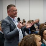 Wśród dużej grupy siedzących osób stoi mężczyzna w okularach. Trzyma w ręce mikrofon.