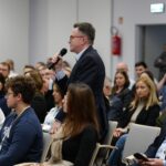 Wśród dużej grupy siedzących osób stoi mężczyzna w okularach. Trzyma mikrofon w ręce.