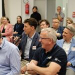 Grupa ludzi siedzi na sali. Wśród nich uśmiechnięty mężczyzna z mikrofonem w ręku.