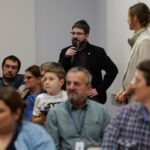 Wśród kilkunastu siedzących osób stoi mężczyzna z brodą, w okularach. Trzyma mikrofon w ręce.