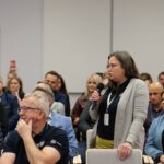 Wśród dużej grupy siedzących osób stoi kobieta w okularach. Mówi do mikrofonu, który trzyma w ręce.