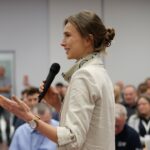 Uśmiechnięta kobieta w jasnym żakiecie mówi do mikrofonu trzymanego w ręku.