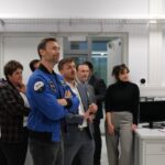 Kilka osób stoi w pomieszczeniu laboratoryjnym, patrząc w jednym kierunku.