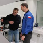 Mężczyzna w niebieskiej kurtce obserwuje, jak mężczyzna w swetrze pokazuje coś przy stanowisku pomiarowym.