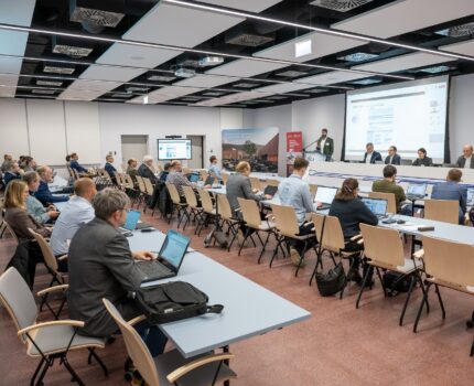 Na sali konferencyjnej, w kilku rzędach, przy stolikach, siedzą uczestnicy spotkania. Na ścianie przed nimi duży ekran z wyświetloną prezentacją.